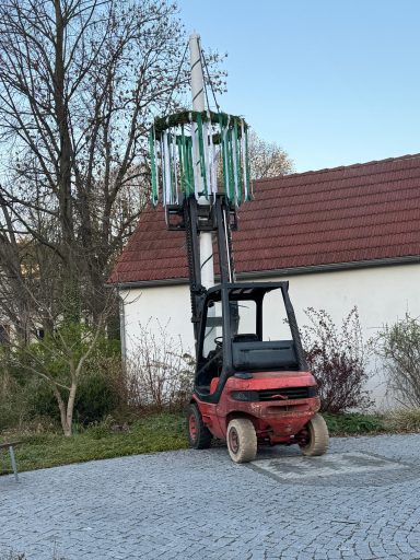 Gabelstapler mit hochgezogenem Aufsatz neben einem Gebäude in einer Gartenlandschaft.