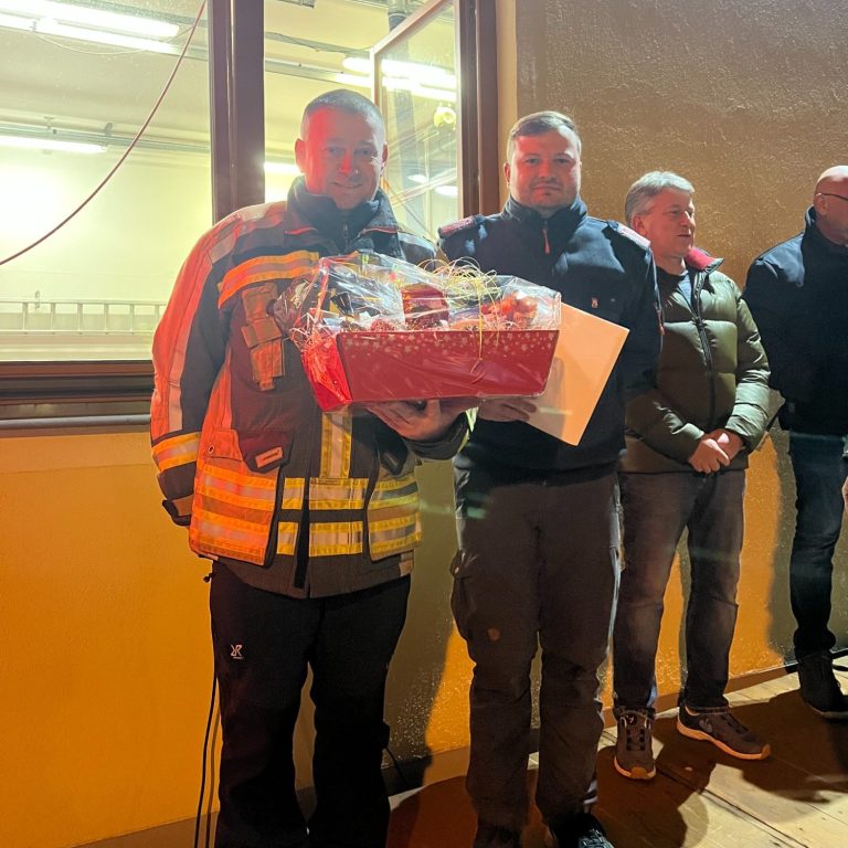 Zwei Personen halten einen Korb mit Geschenken, im Hintergrund stehen weitere Personen.