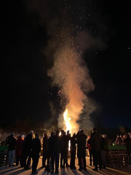 Menschen stehen vor einem hohen Feuer in der Dunkelheit, Funken fliegen nach oben.