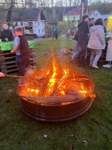 Feuerstelle mit brennendem Holz, umgeben von Menschen und Gebäuden im Hintergrund.