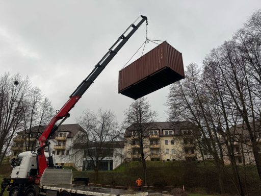 Ein Kran hebt einen Container über ein Gelände mit Wohngebäuden im Hintergrund.