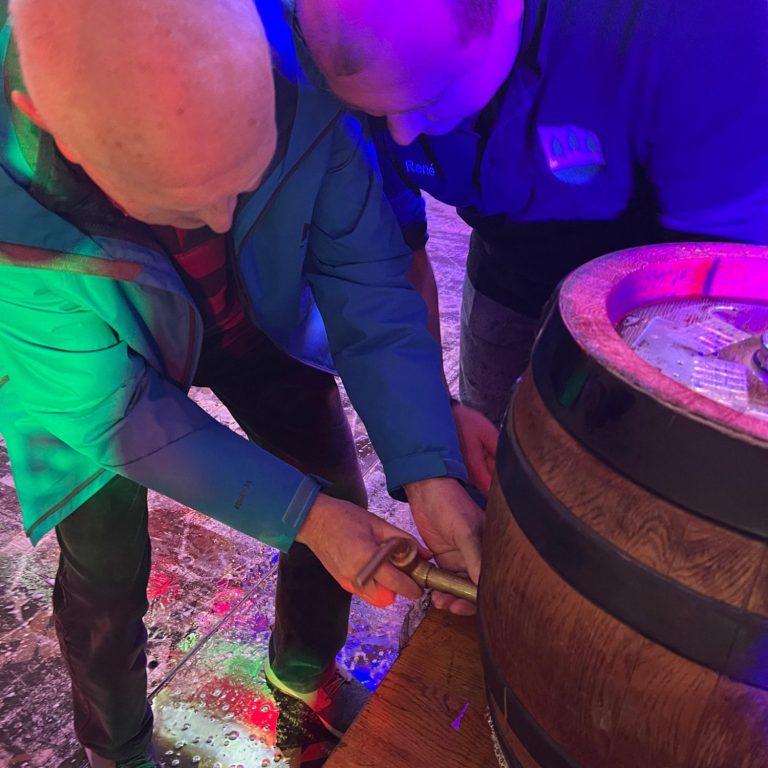 Zwei Personen arbeiten gemeinsam an einem Holzfass. Bunte Lichter im Hintergrund.