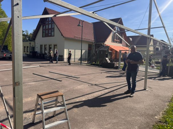 Aufbau eines Rahmens vor einem Gebäude mit Personen und einer Leiter im Vordergrund.