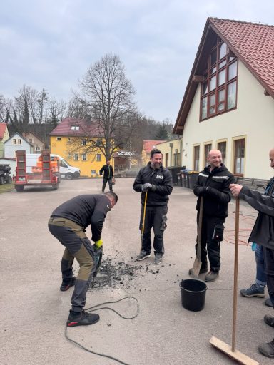 Männer arbeiten auf einer Baustelle, einige halten Werkzeuge, ein Mann gräbt im Boden.