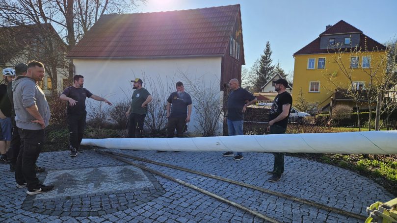 Gruppierung von Menschen, die eine lange, weiße Stange auf einem gepflasterten Platz halten.