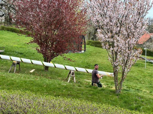 Mann arbeitet an einer langen Röhre in einer grünen Landschaft mit blühenden Bäumen.
