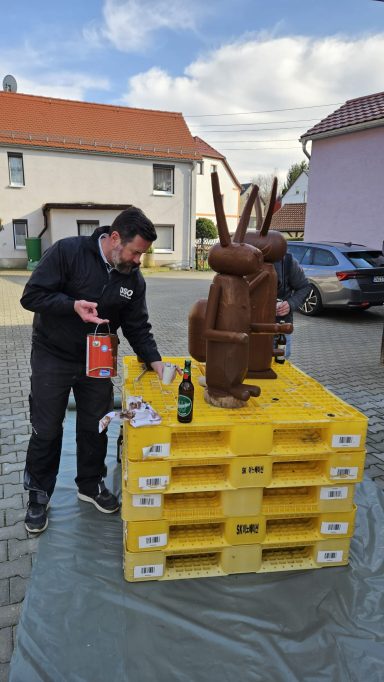 Person arbeitet an einer kreativen Skulptur aus Holz, umgeben von Paletten.