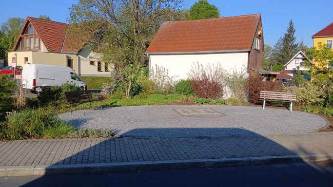 Ein ruhiger Platz mit Häusern, Bäumen und einer Bank in der Nähe.