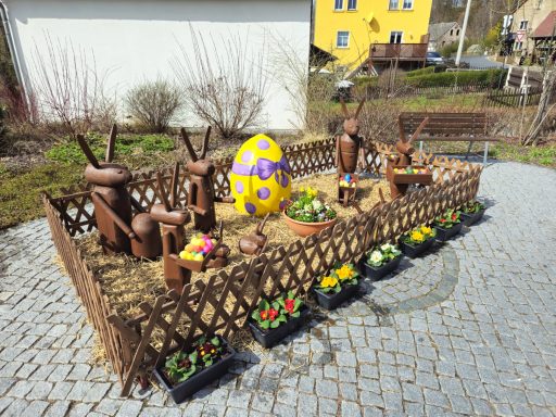 Osterdekoration mit einem großen Ei und mehreren Figuren aus Holz in einem Garten.