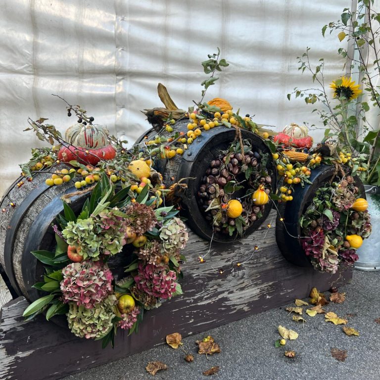 Drei dekorierte Fässer mit bunten Blumen und Früchten, umgeben von herbstlichem Laub.
