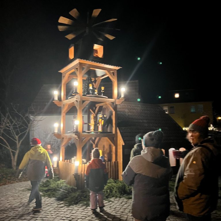 Weihnachtliche Holzpyramide mit Lichtern, umgeben von Menschen bei Nacht.