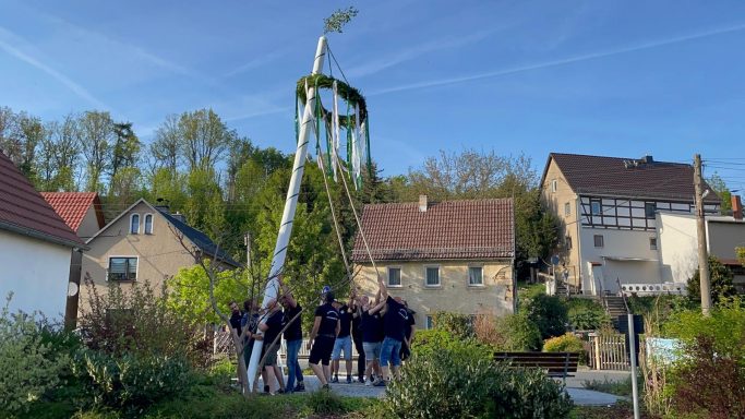 Menschen versammeln sich um einen großen Maibaum in einer ländlichen Umgebung.