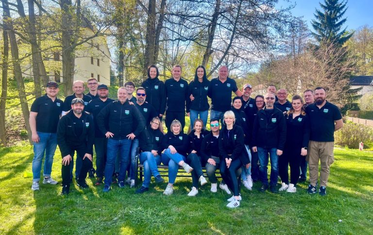 Gruppe von 25 Personen in schwarzen T-Shirts, stehend im Freien zwischen Bäumen.