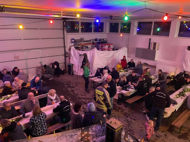 Menschen sitzen in einer festlich dekorierten Halle mit Tischen und bunten Lichtern.